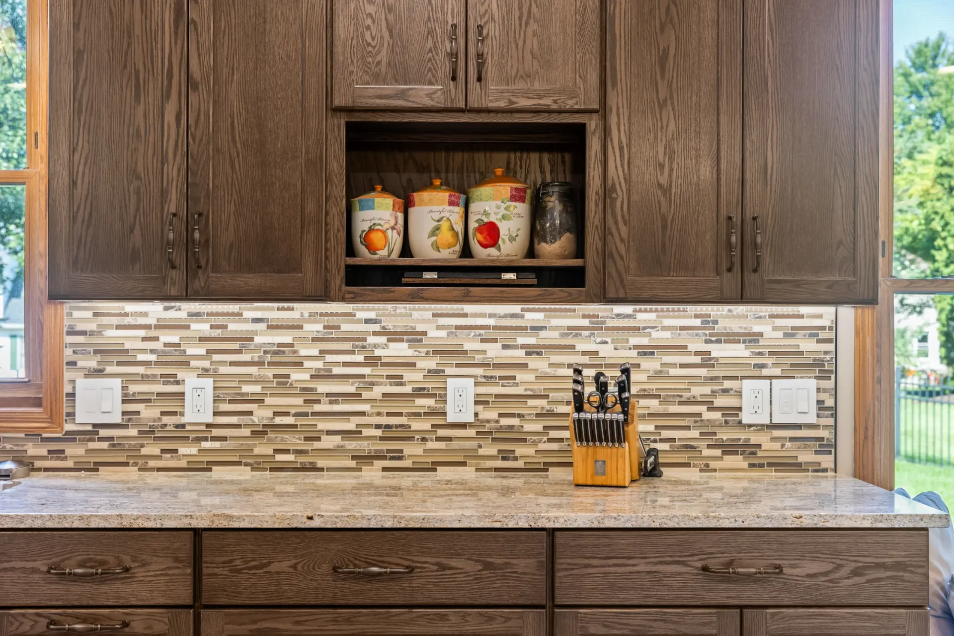 Modern kitchen counter with a knife block, mosaic tile backsplash, and dark wood cabinets. Three decorative canisters are displayed on an open shelf above the counter.