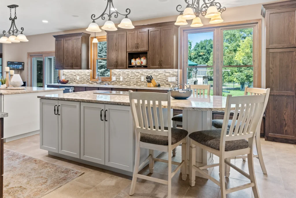 Modern kitchen with a large island, light granite countertops, wooden cabinets, pendant lighting, four chairs, and large windows overlooking a green backyard.