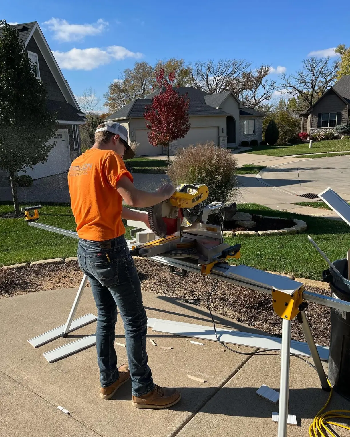 A person in an orange shirt operates a miter saw to cut white boards on a driveway in a suburban neighborhood.