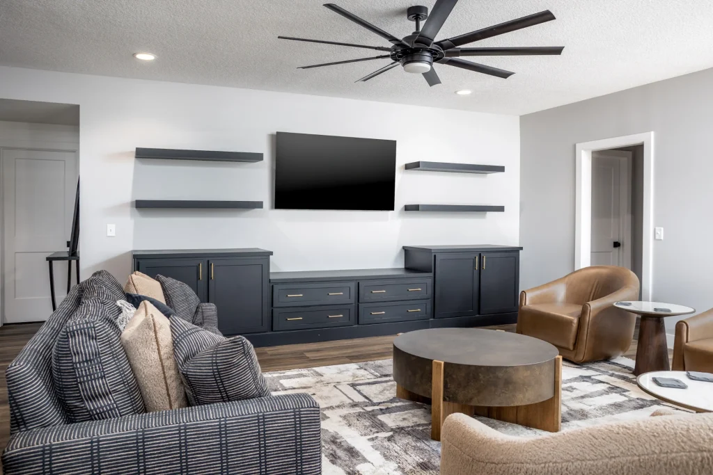 Modern living room with a black entertainment unit, wall-mounted TV, floating shelves, round coffee table, armchairs, and neutral-toned decor—designed for custom comfort beyond builder grade standards.