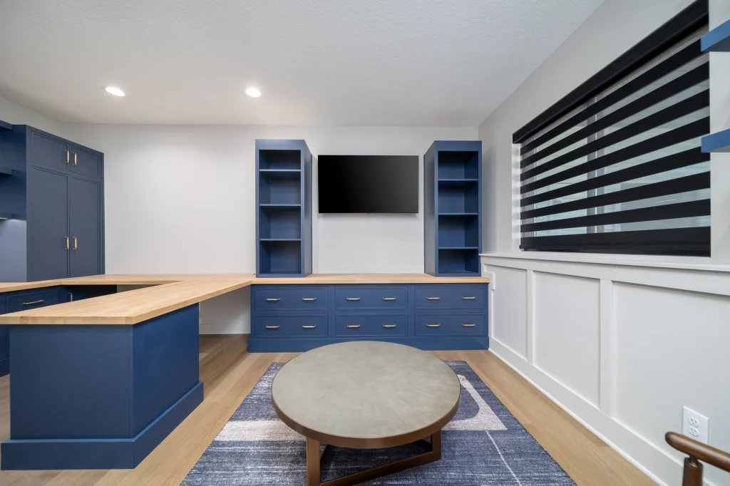 Modern home office with blue cabinetry, built-in shelves, a wall-mounted TV, wooden desk, round table, and a window with striped blinds—offering custom comfort far beyond builder grade finishes.