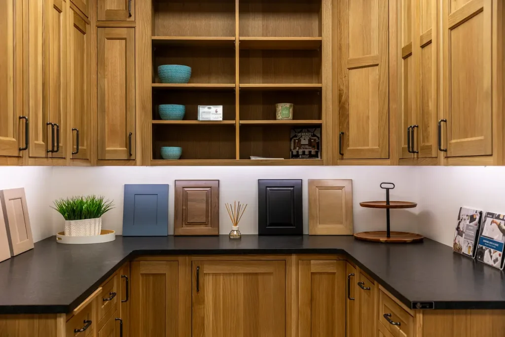 A corner display in the showroom features wooden kitchen cabinets with sample cabinet doors, decorative items, and shelving above a black countertop.