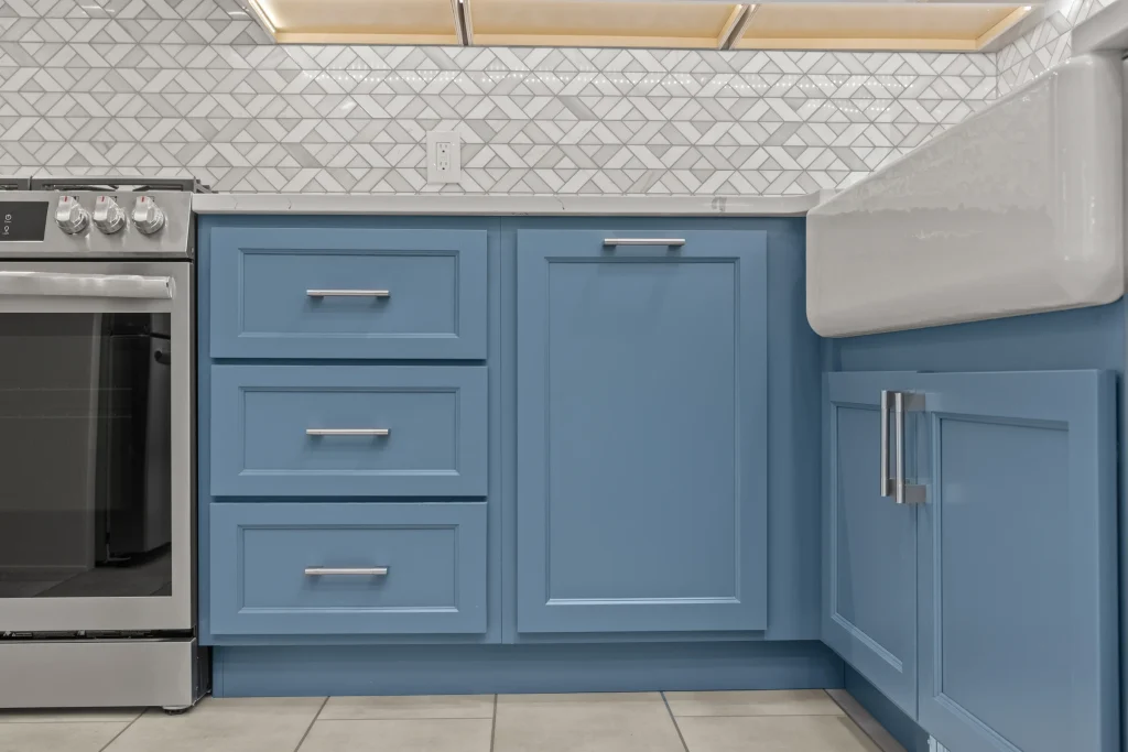Blue kitchen cabinets with silver handles, a white farmhouse sink, geometric white tile backsplash, and part of a stainless steel stove visible on the left.