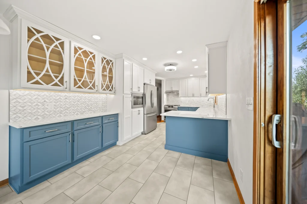 Modern kitchen with blue lower cabinets, white upper cabinets with glass doors, stainless steel appliances, light tile flooring, and a patterned white backsplash.