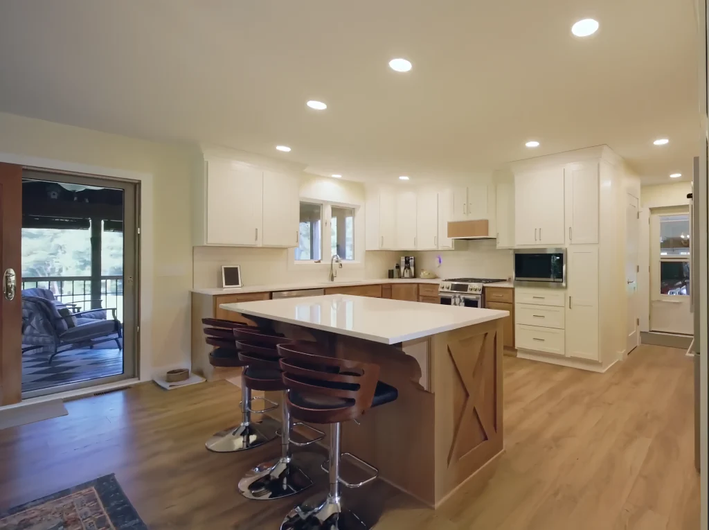 Modern kitchen remodel in Milan, IL with white cabinets, a central island with wooden base, three bar stools, stainless steel appliances, and wood flooring; sliding glass door opens to a porch.