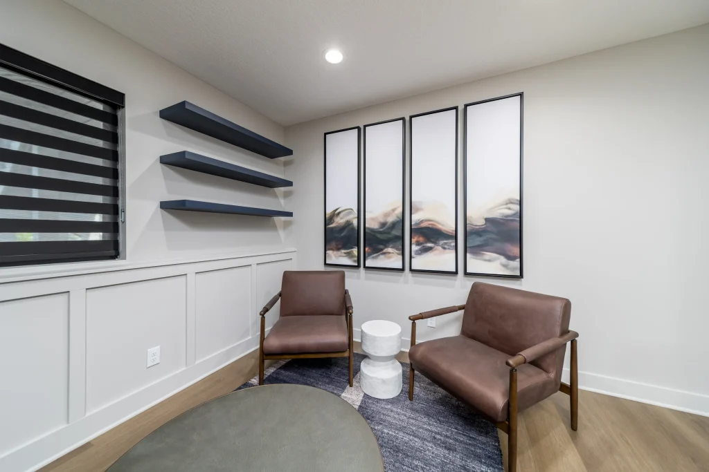 A modern sitting area with two brown chairs offering custom comfort, a round table, three floating shelves, and four abstract art panels on a light-colored wall.