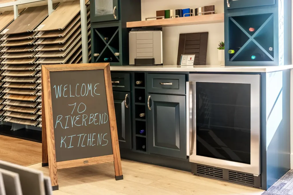 A chalkboard sign reads “Welcome to Riverbend Kitchens” in front of a modern showroom display featuring sample cabinets and appliances.