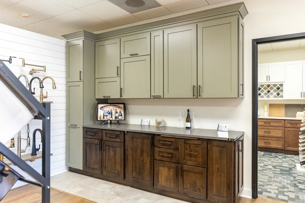 Discover our modern kitchen Showroom, featuring tall green upper cabinets, dark wood lowers, a gray countertop, wine bottles, and a small TV—an inspiring display on our Showroom Page.