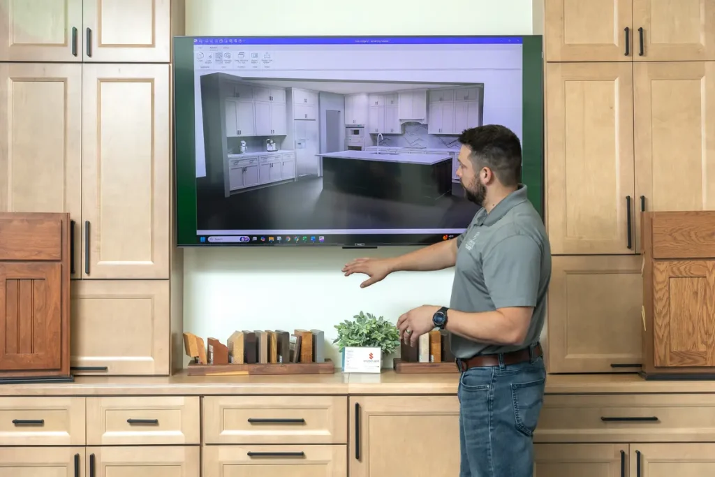 A man stands in front of wooden cabinets in a showroom, pointing at a large screen displaying a kitchen design rendering.