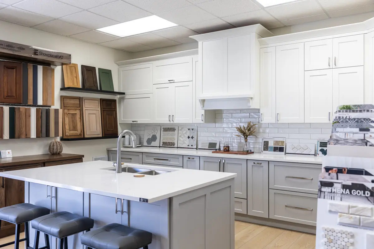 A modern kitchen showroom page featuring white cabinets, a white island with gray stools, and displays of tile and wood cabinet door samples on the walls and counters.
