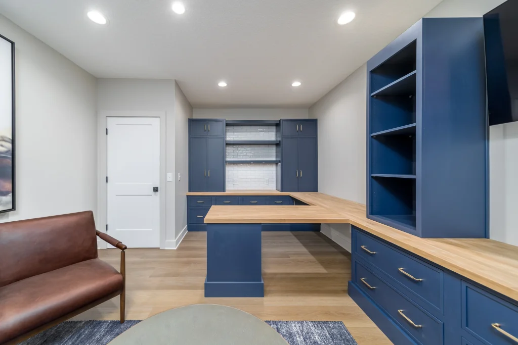 Modern home office with blue cabinetry, wooden countertops, custom comfort seating in a brown leather chair, open shelves, and wood flooring under recessed lighting—far beyond builder grade finishes.