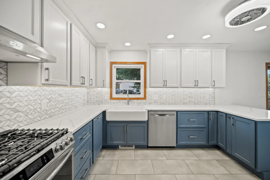 Modern kitchen with blue lower cabinets, white upper cabinets, a farmhouse sink, stainless steel appliances, geometric tile backsplash, and a window above the sink.
