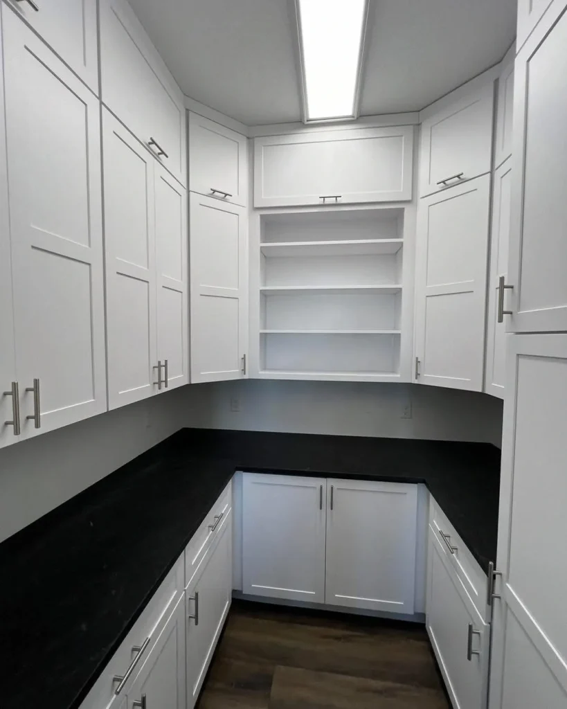 A pantry room with builder grade white cabinets, black countertops, open shelves in the center, and wood flooring under overhead fluorescent lighting offers custom comfort and functionality.