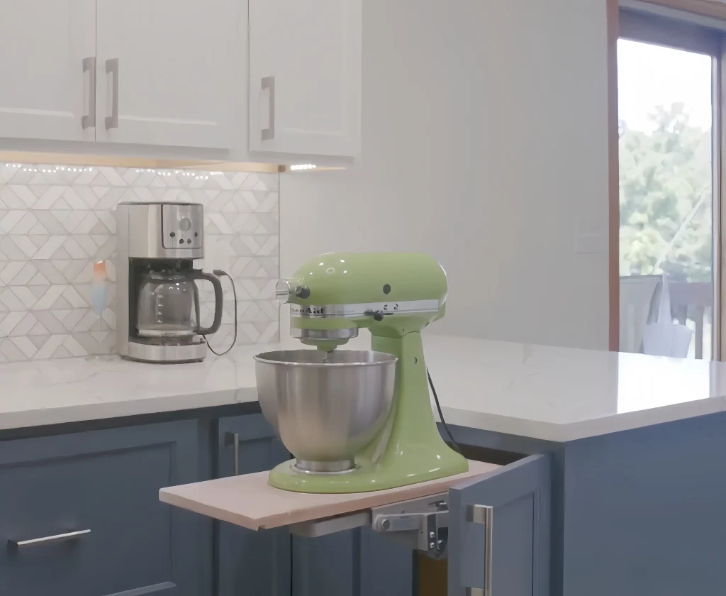 A green stand mixer is placed on an extended pull-out shelf in a modern kitchen with blue cabinets and white countertops. A coffee maker sits on the counter in the background.