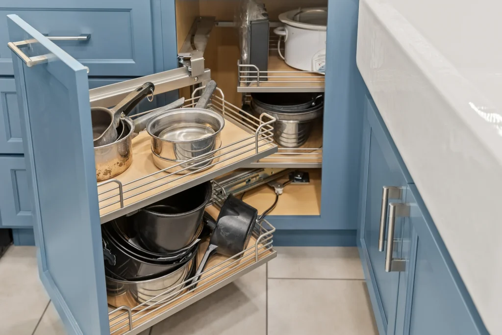 A kitchen cabinet with blue doors is open, revealing pull-out metal shelves filled with pots, pans, and kitchenware.