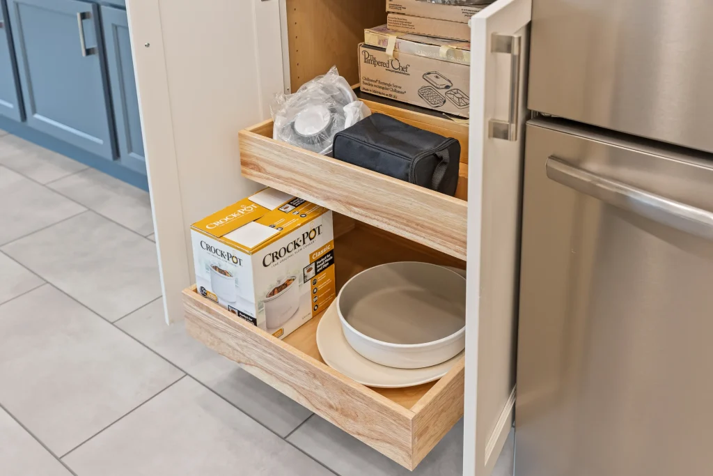 Open kitchen cabinet with wooden pull-out shelves holding Crock-Pot boxes, plastic-wrapped items, a folded cloth, and stacked white plates, next to a stainless steel refrigerator.