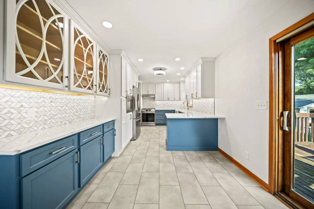Modern kitchen with blue lower cabinets, white upper cabinets, stainless steel appliances, patterned backsplash, tile flooring, and a sliding glass door leading to a wooden deck.