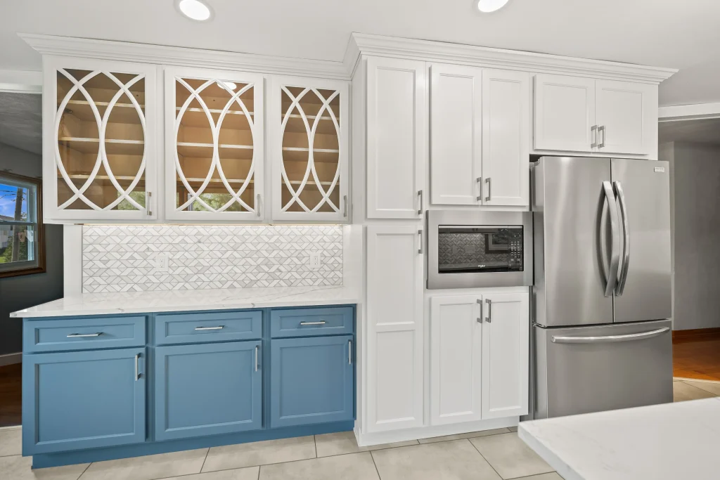 Modern kitchen with white upper cabinets featuring glass doors, blue lower cabinets, stainless steel refrigerator, built-in microwave, and white patterned backsplash.