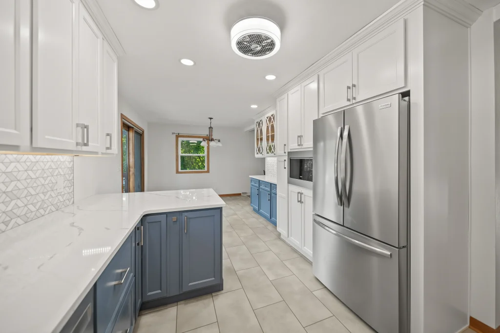 Modern kitchen with white upper cabinets, blue lower cabinets, stainless steel appliances, tile floor, and a window overlooking greenery.
