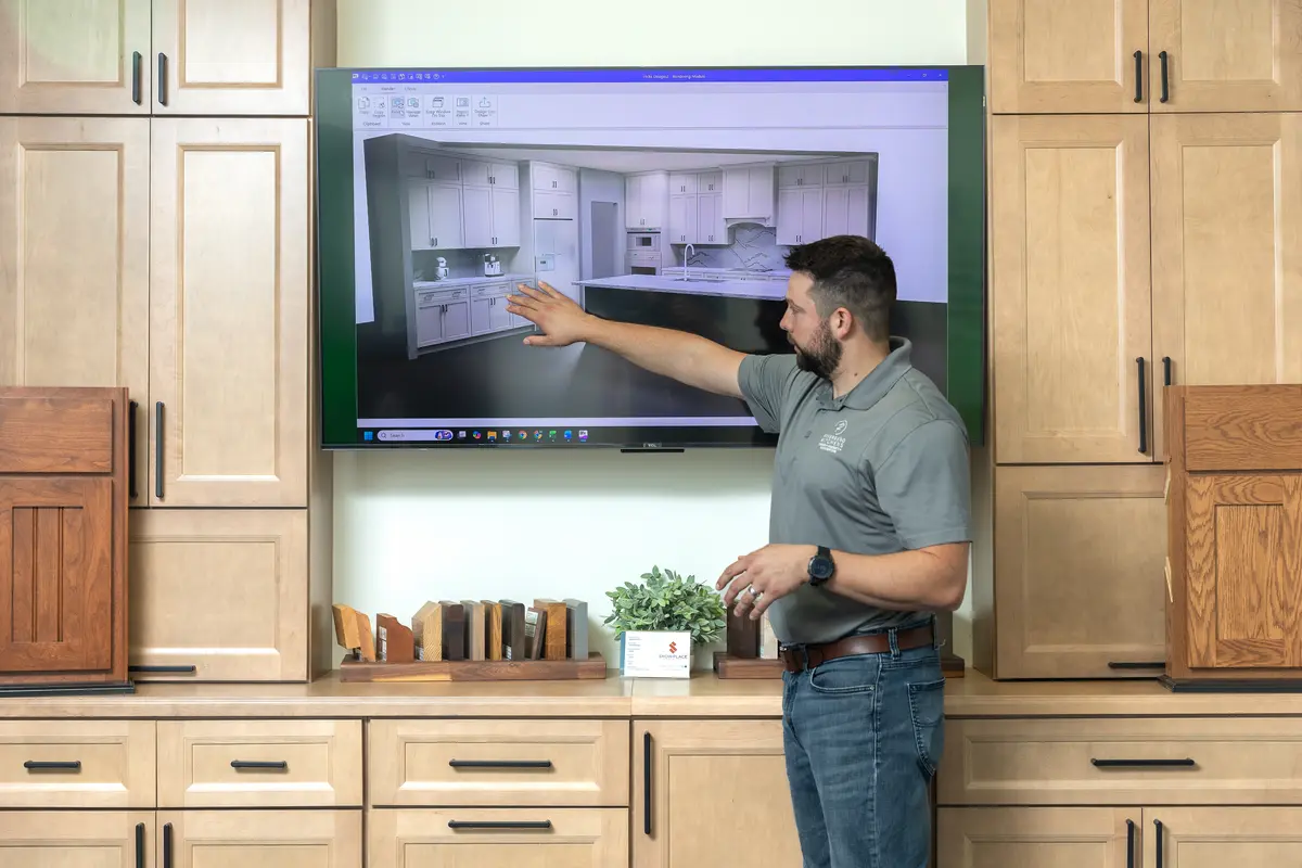 A man points to a kitchen design displayed on a large screen in a showroom, standing in front of wooden cabinets and countertop material samples.