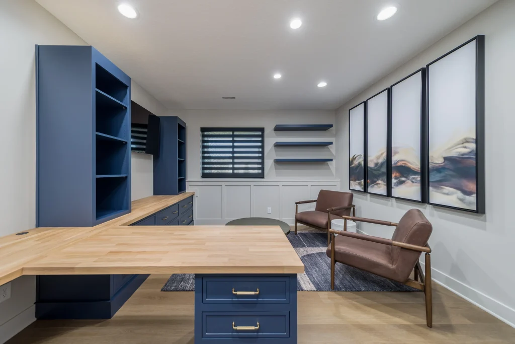 Modern home office with blue built-in shelves, wooden desk, two brown chairs, abstract wall art, and recessed lighting on the ceiling—designed for custom comfort beyond builder grade standards.
