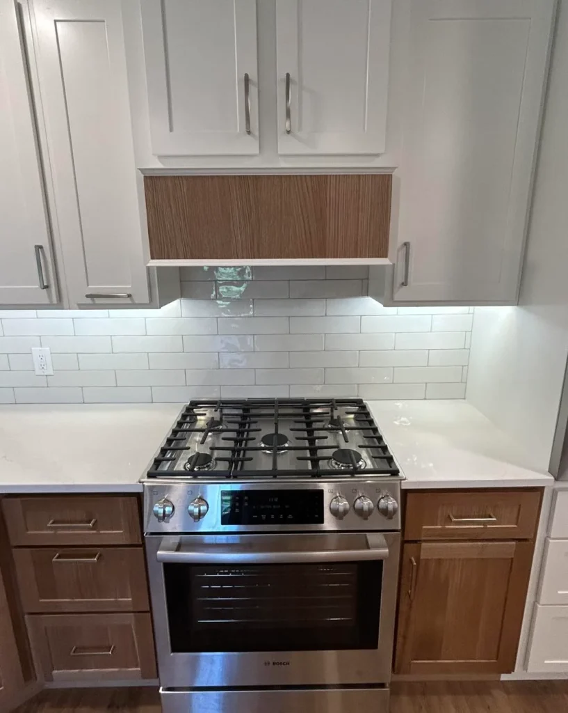 A stainless steel gas stove with an oven is set between white countertops and wood cabinets in this Milan IL kitchen remodel, featuring a white tile backsplash and upper white cabinets above.