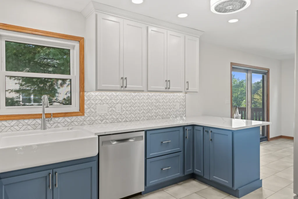 Modern kitchen with white upper cabinets, blue lower cabinets, a farmhouse sink, stainless steel dishwasher, geometric backsplash, and large window and glass door with outdoor view.