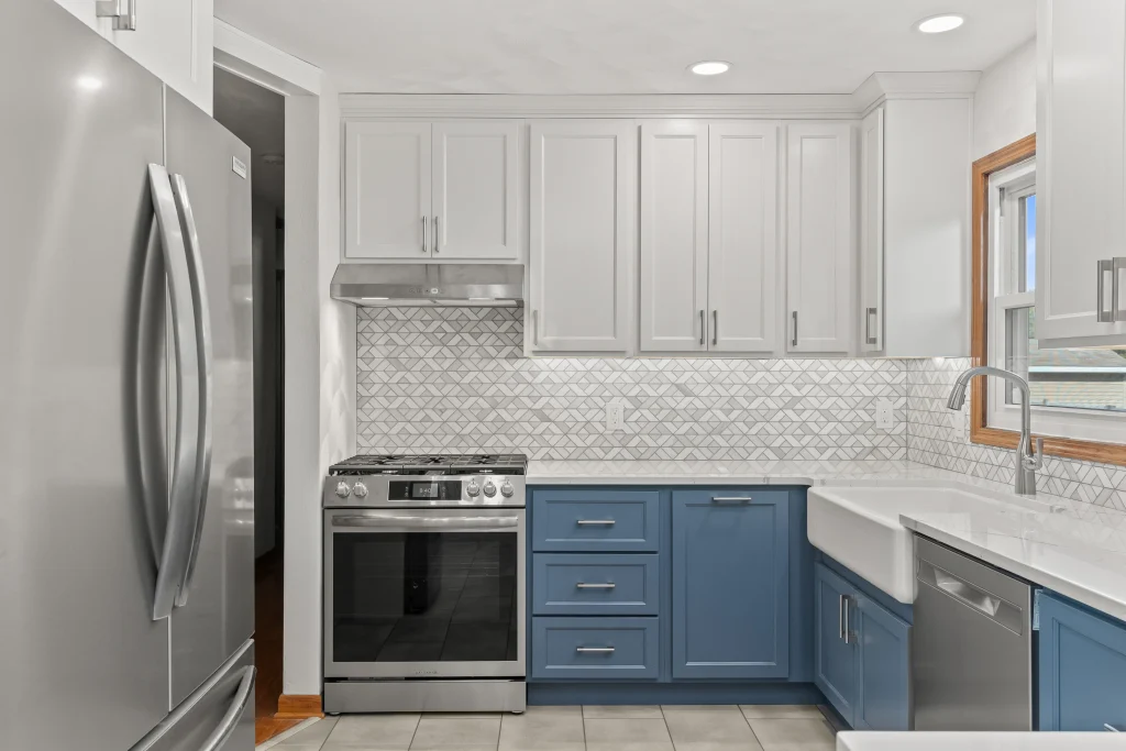 Modern kitchen with stainless steel appliances, white upper cabinets, blue lower cabinets, geometric tile backsplash, and a farmhouse sink by a window.