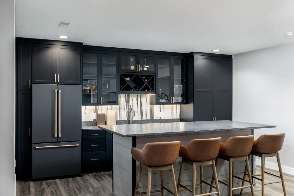 Featured Project: Modern kitchen with dark cabinets, a gray island countertop, brown leather bar stools, and built-in appliances. Marble backsplash and open shelving complete the sophisticated look above the sink area.