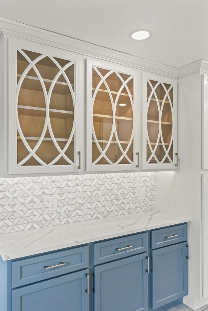 Upper cabinets with glass doors featuring geometric white trim, blue lower cabinets, marble countertop, and a patterned tile backsplash in a kitchen.