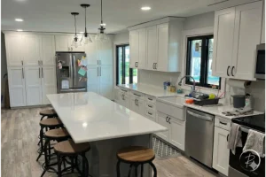 Modern kitchen with white cabinets, a long marble island with stools, stainless steel appliances, and large windows above the sink.