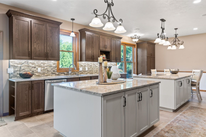 Modern kitchen with two-tone cabinetry, granite countertops, pendant lighting, stainless steel appliances, and a large island with decor items. Dining area visible in the background.