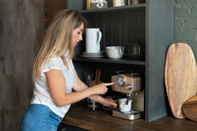 A woman uses an espresso machine to make coffee in a kitchen with shelves holding dishes and kitchenware.