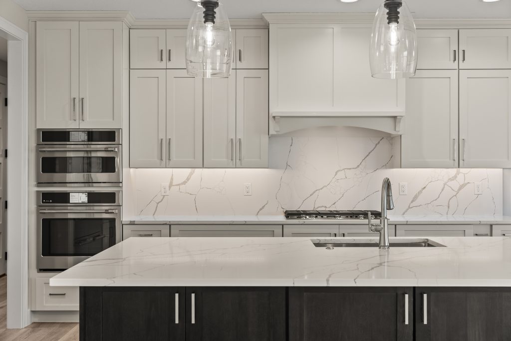 Modern white kitchen with marble backsplash, stainless steel double ovens, and dark island cabinets with a marble countertop; two glass pendant lights above.