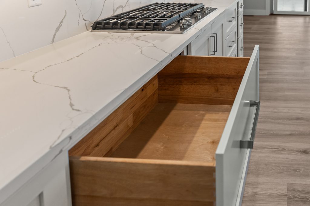 Open drawer under a white marble countertop with a gas cooktop above in a modern gray-kitchen setting, showing wood interior and soft gray cabinets.