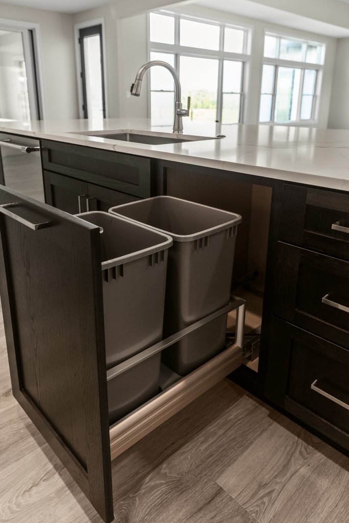 Open pull-out drawer revealing two gray trash/recycling bins under a modern kitchen island with a marble countertop and dark wood cabinetry.