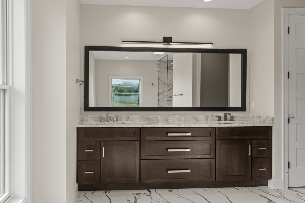 Wide bathroom vanity with dark wood drawers, marble countertop, and a large framed mirror; horizontal light fixture above the mirror appears in view.