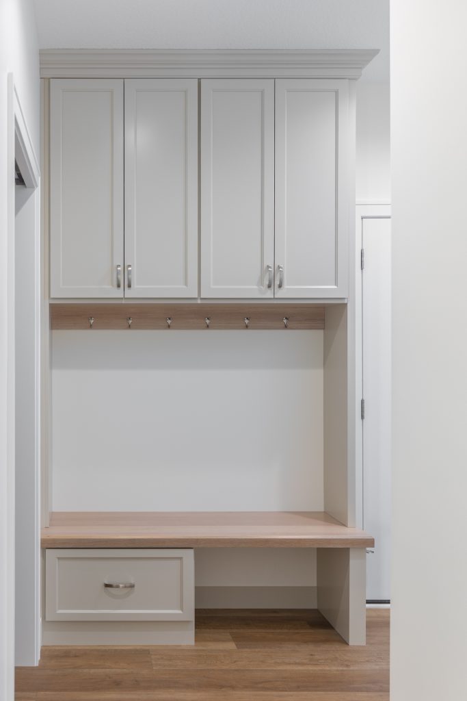 White built-in storage unit with upper cabinet doors, a wooden peg rail, and a lower desk-like shelf in a narrow hallway area