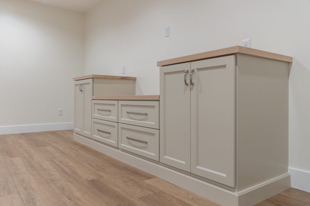 Corner view of beige kitchen cabinets with light wood countertops and a wooden floor in a bright room.
