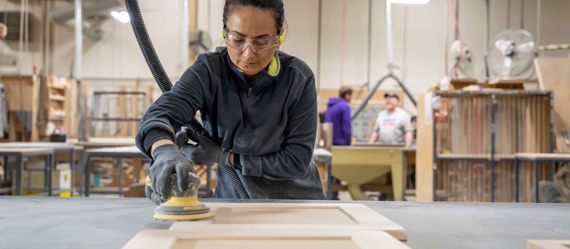 Sanding2-1830x800-1 Worker wearing safety goggles and gloves sands a wooden panel on a table in a woodworking shop.
