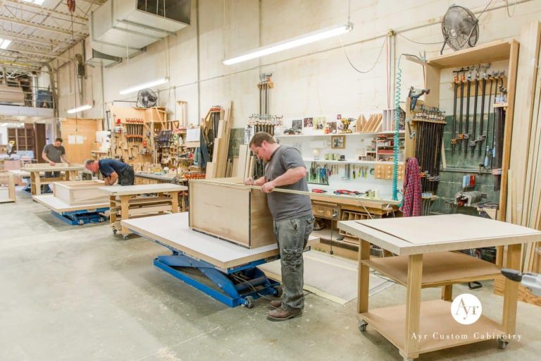Carpenters building and assembling cabinets in a busy woodshop with clamps, sawdust, and workbenches; AyR Custom Cabinetry logo visible in the corner.